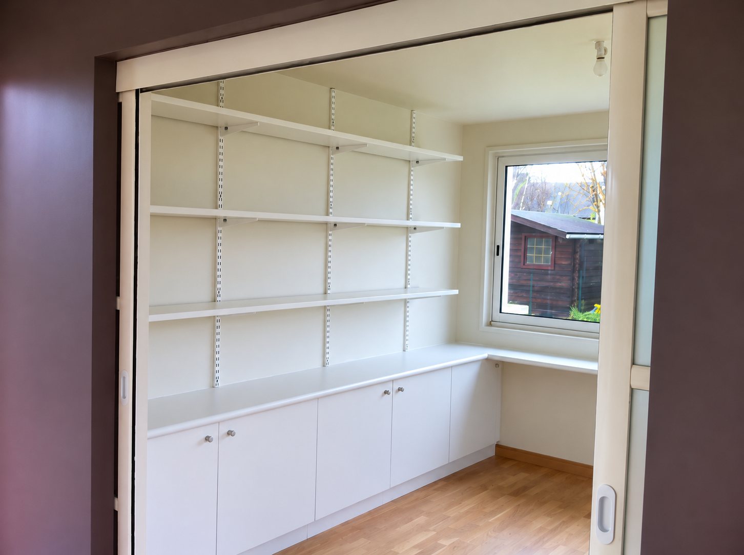 Une petite pièce au parquet clair équipée d'étagères murales blanches sur crémaillères et de placards bas blancs, avec une fenêtre donnant sur un abri de jardin.