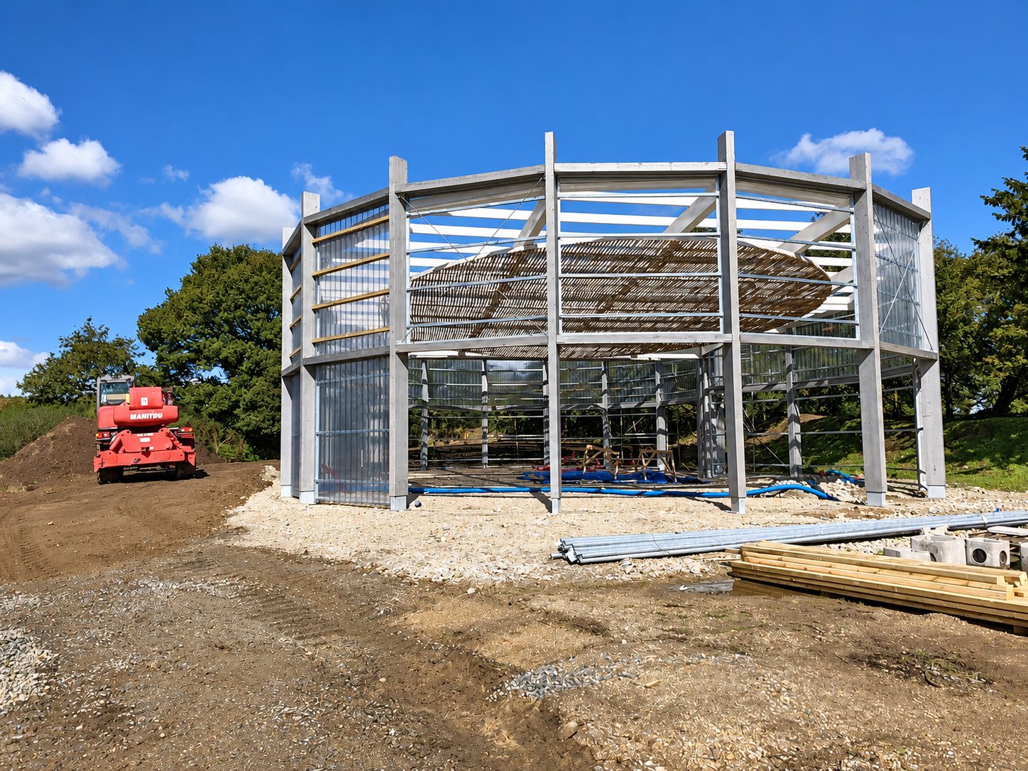 Halles de Lanrivain. Structure métallique polygonale en construction sur un terrain de gravier, avec un chariot télescopique Manitou rouge à gauche et des matériaux au premier plan.