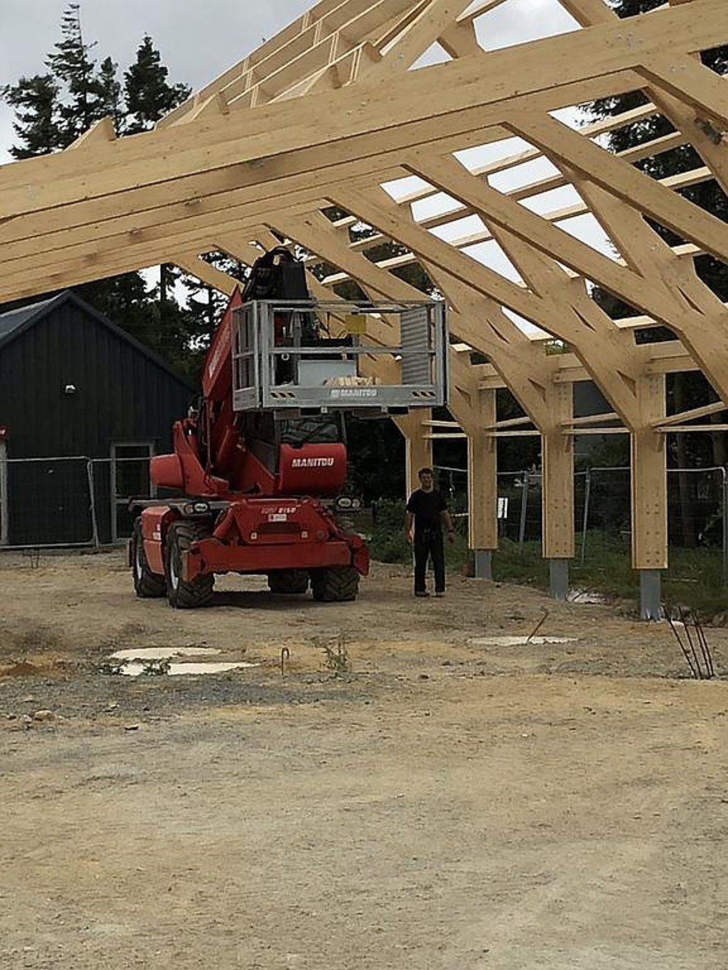 Sur un chantier, un télescopique Manitou rouge avec nacelle se tient sous une grande charpente en bois, près d'un homme debout.