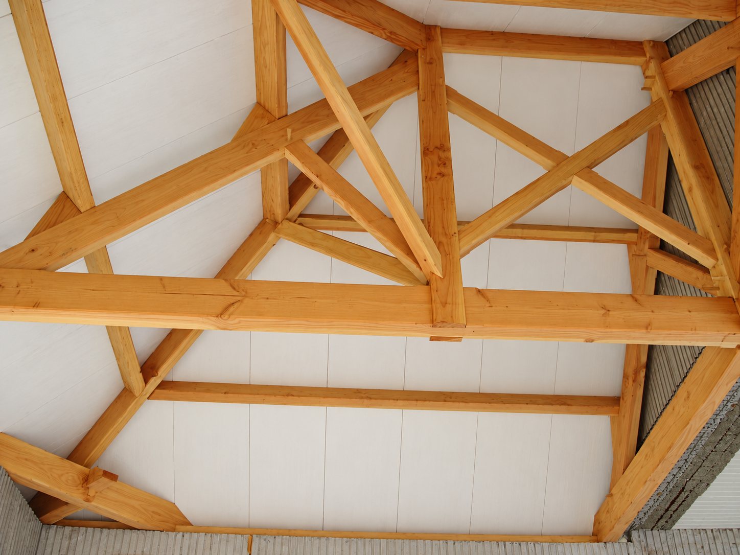 Vue de dessous d'une charpente traditionnelle en bois clair assemblée sous un plafond de panneaux blancs, dans une construction en cours.