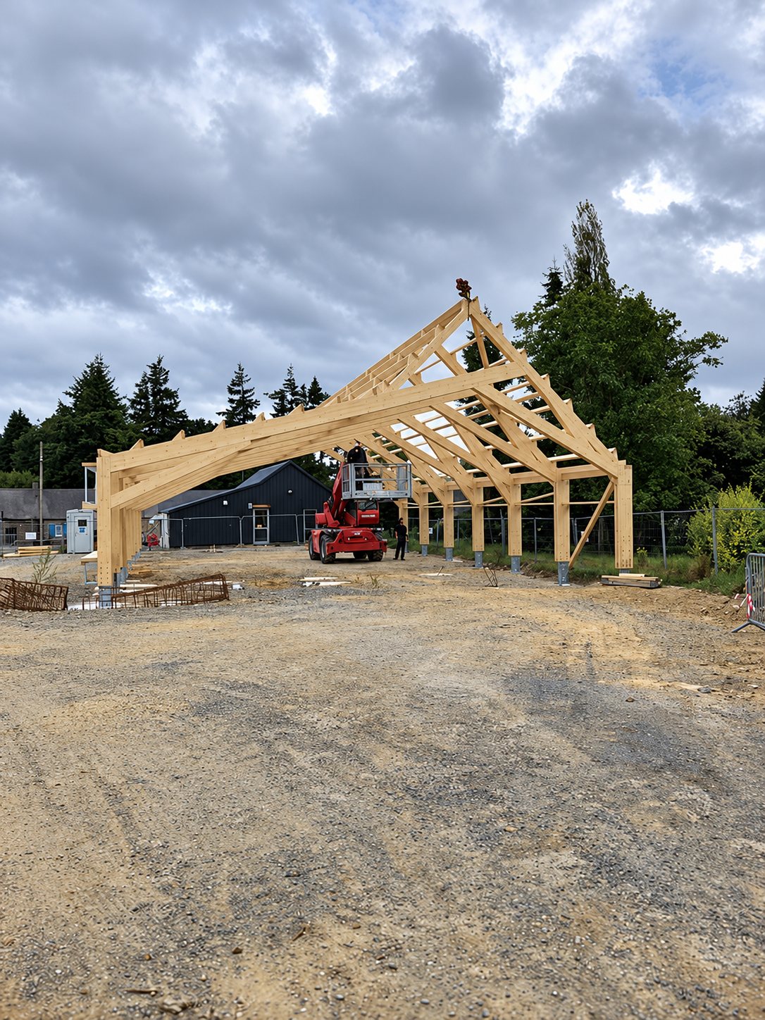 Boulodrome de Moustéru. Une grande charpente en bois clair est dressée sur un chantier en terre, avec un engin télescopique rouge à l'arrière et des arbres sous un ciel nuageux.
