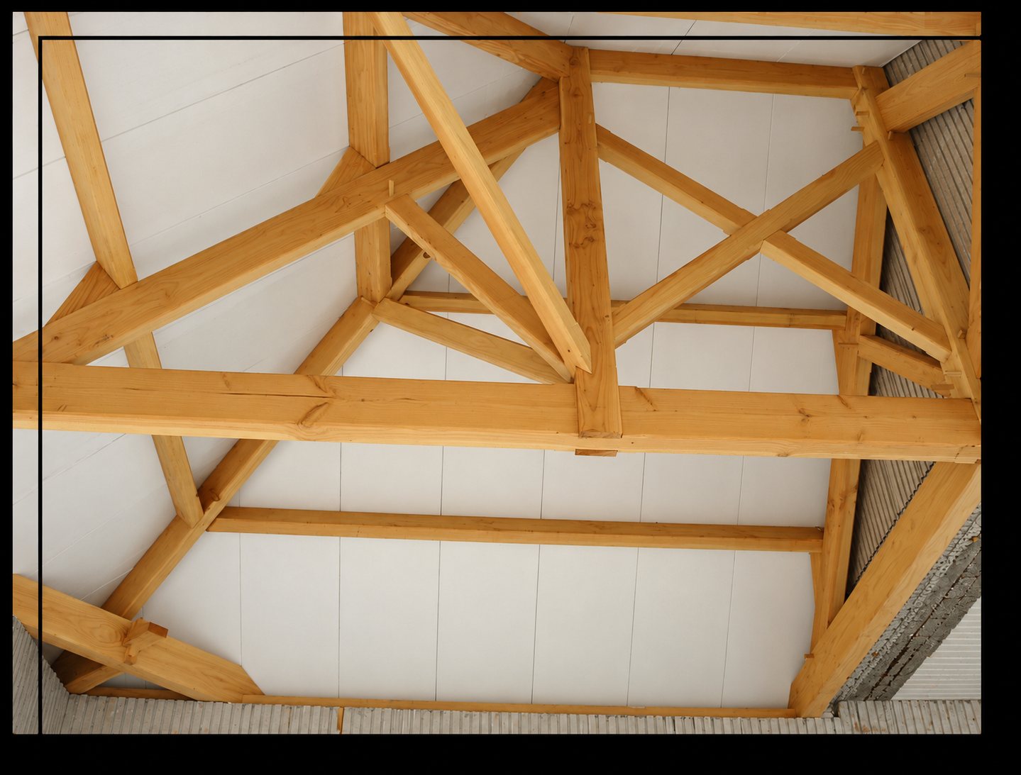 Vue de dessous d'une charpente traditionnelle en bois clair, avec poutres et jambes de force, installée sous un plafond blanc dans une construction en cours.