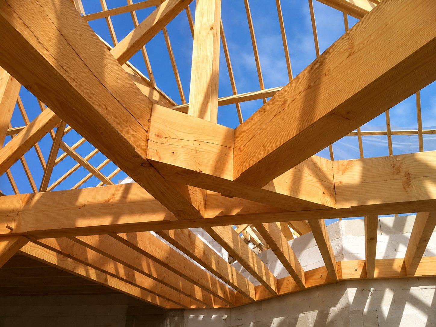 Vue de l'intérieur d'une charpente en bois clair en cours de montage, avec des poutres et chevrons se détachant sur un ciel bleu, au-dessus de murs en béton cellulaire.