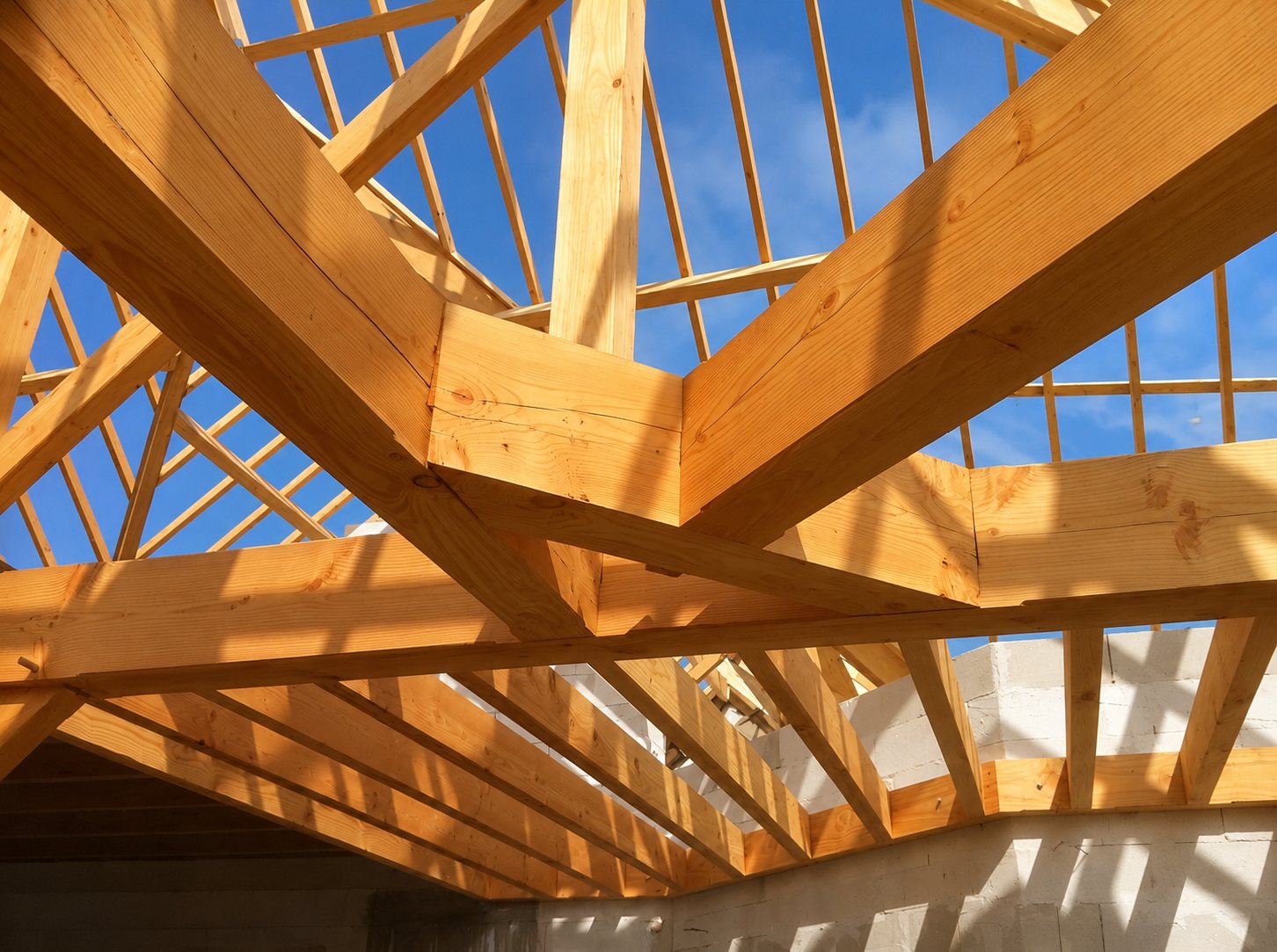 Maisons Perros Guirec. Vue depuis l'intérieur d'une charpente en bois clair en cours de montage, avec des poutres et des chevrons sous un ciel bleu, au-dessus de murs en béton cellulaire.