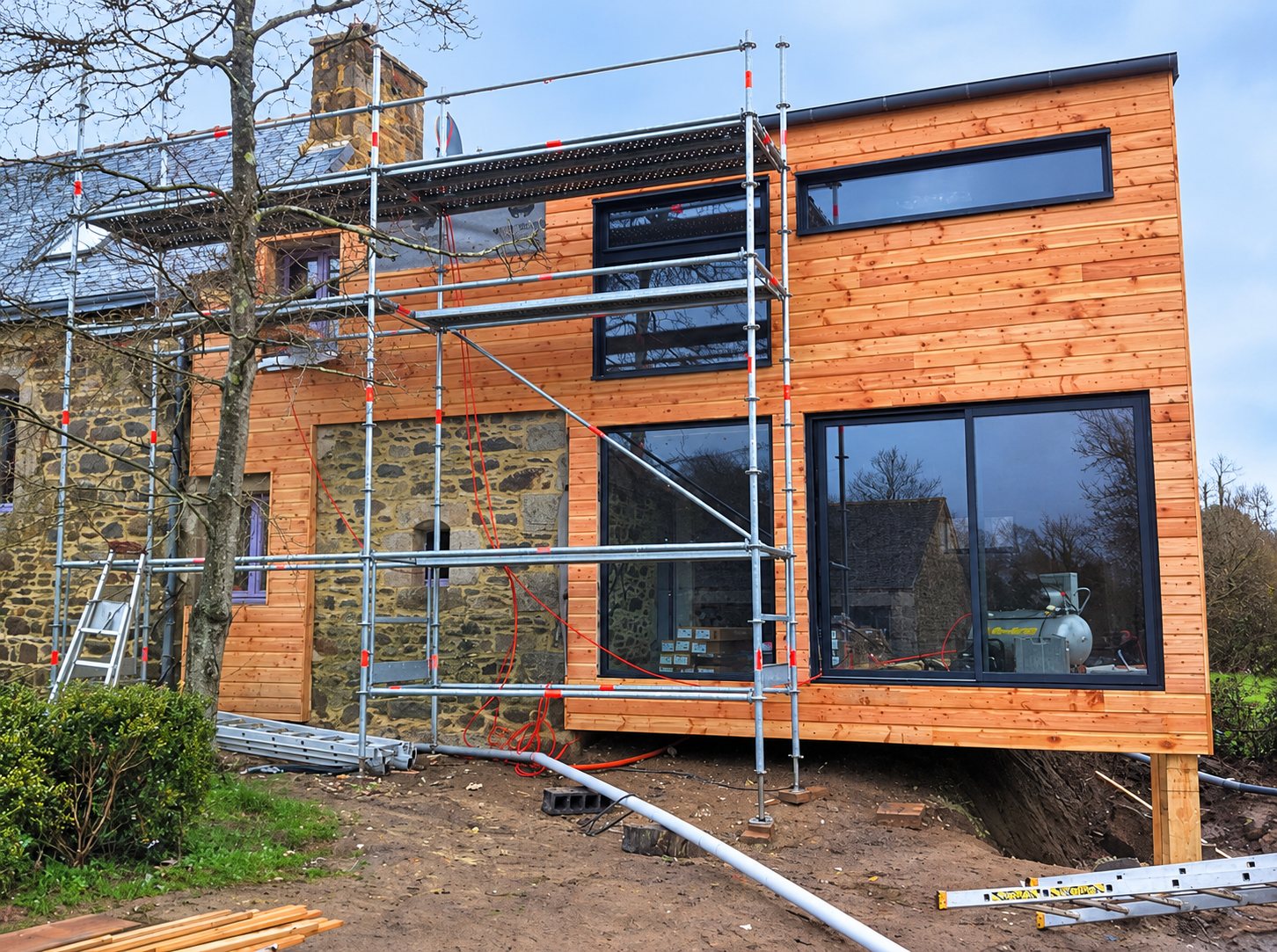 Maison en bois et terrasse. Chantier d'une extension à bardage en bois et grandes baies vitrées noires, accolée à une maison en pierre et entourée d'un échafaudage.