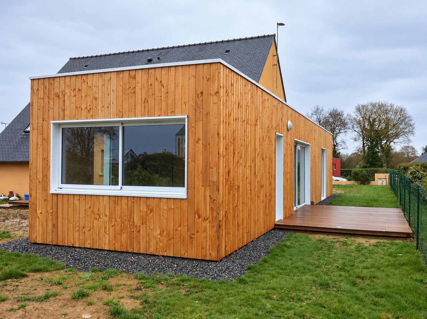 Ossature en bois. Extension de maison à toit plat habillée de bardage bois vertical, avec grande fenêtre coulissante blanche et terrasse en bois donnant sur un jardin.