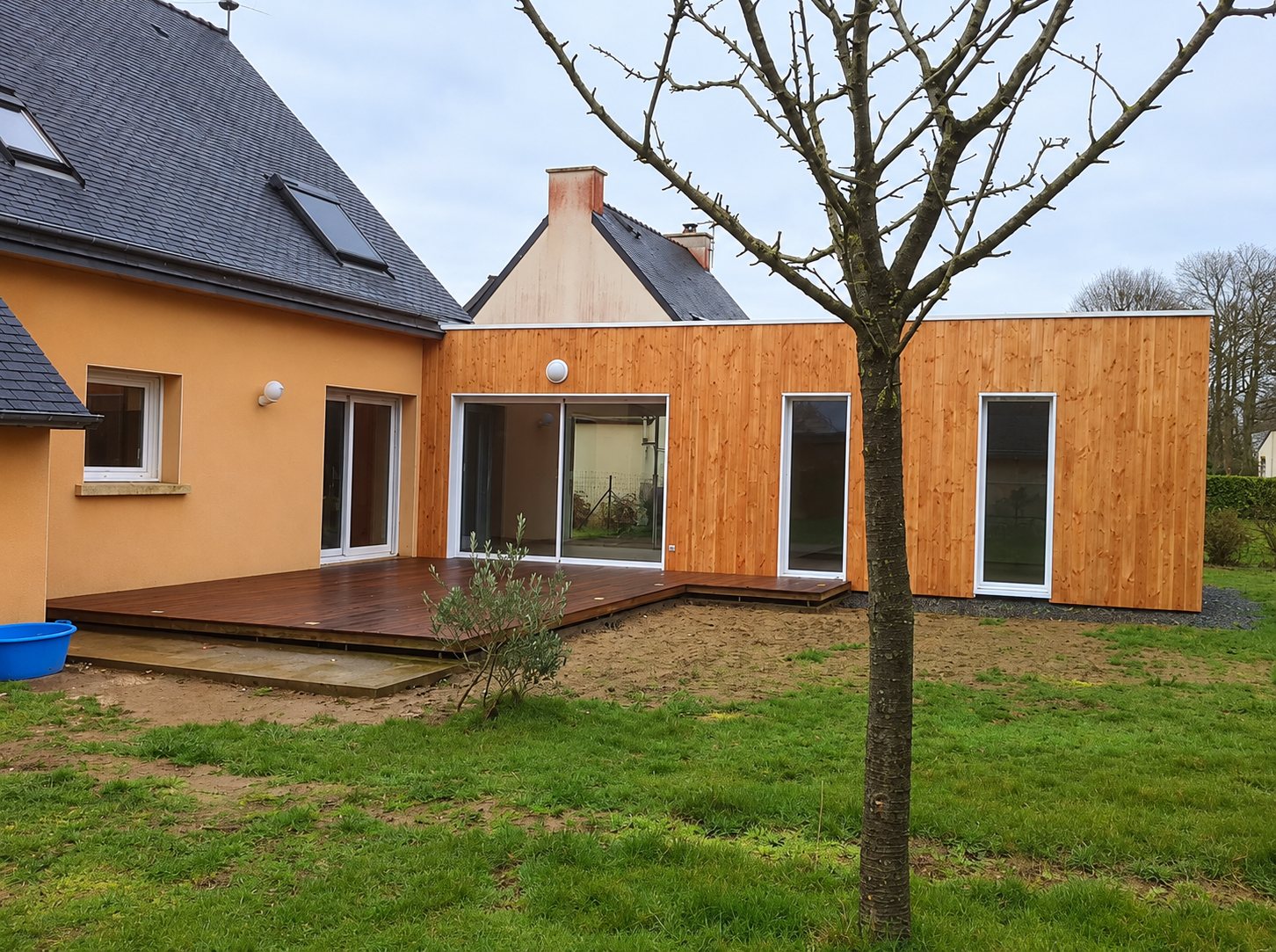 Une extension moderne à bardage en bois claire est accolée à une maison orangée à toit d'ardoise, avec une terrasse en bois et un jardin au premier plan.