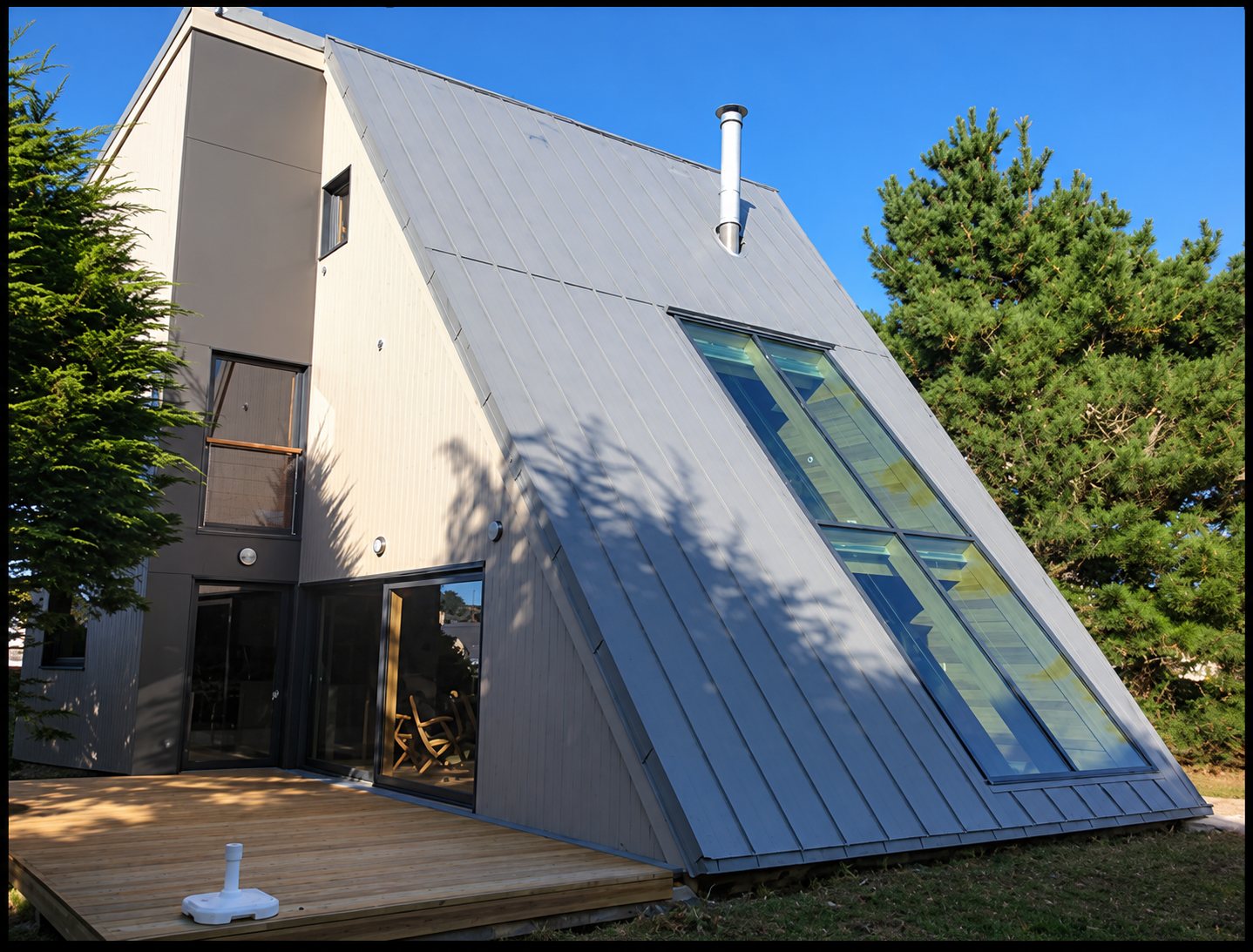 Maison moderne de forme triangulaire dont le grand pan de toiture en zinc est percé d'une verrière, avec une terrasse en bois et des pins autour.