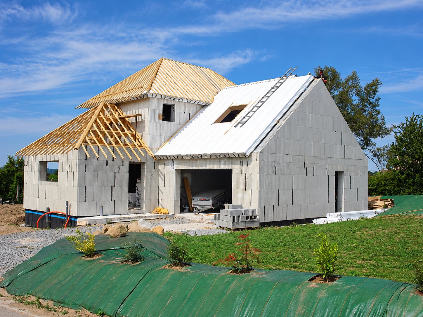 Chantier d'une maison aux murs en blocs isolants, avec une charpente en bois partiellement posée et un toit recouvert de panneaux blancs, sous un ciel bleu.