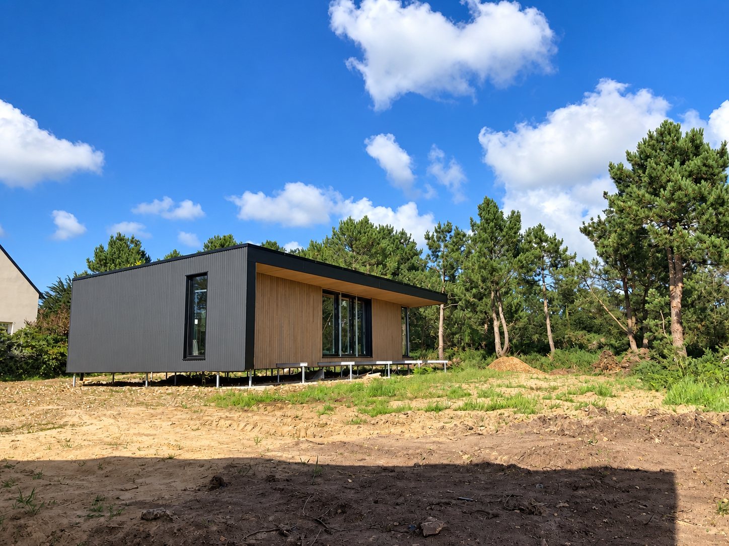 Une maison contemporaine à bardage noir et bois, posée sur des pilotis métalliques dans un terrain sableux entouré de pins sous un ciel bleu.