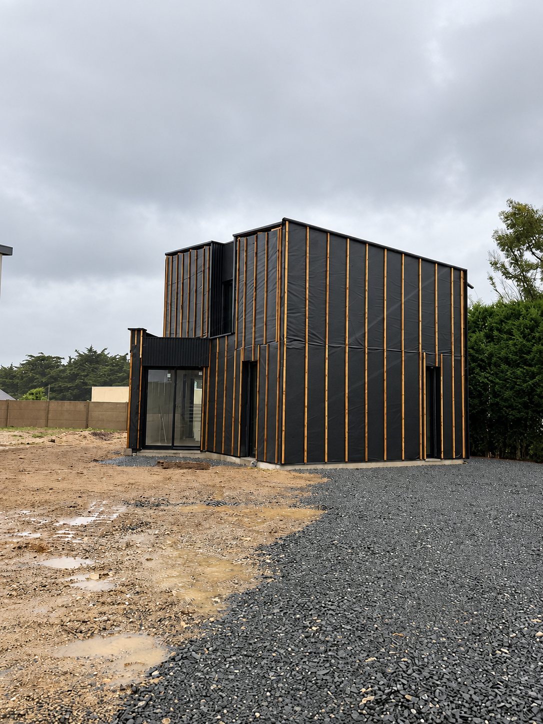 Une maison à étage de forme cubique, en cours de construction, recouverte d'un pare-pluie noir fixé par des tasseaux de bois, sur un terrain de terre et de gravier par temps gris.