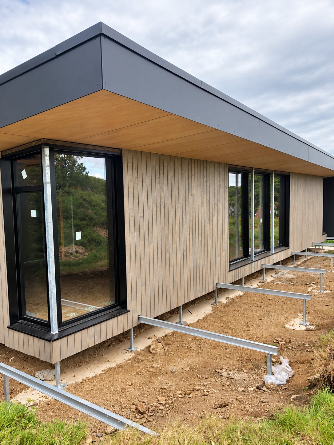 Construction maison individuelle. Façade d'une maison moderne en construction avec bardage bois vertical, grandes fenêtres noires, toit plat à débord et terrain en terre devant.