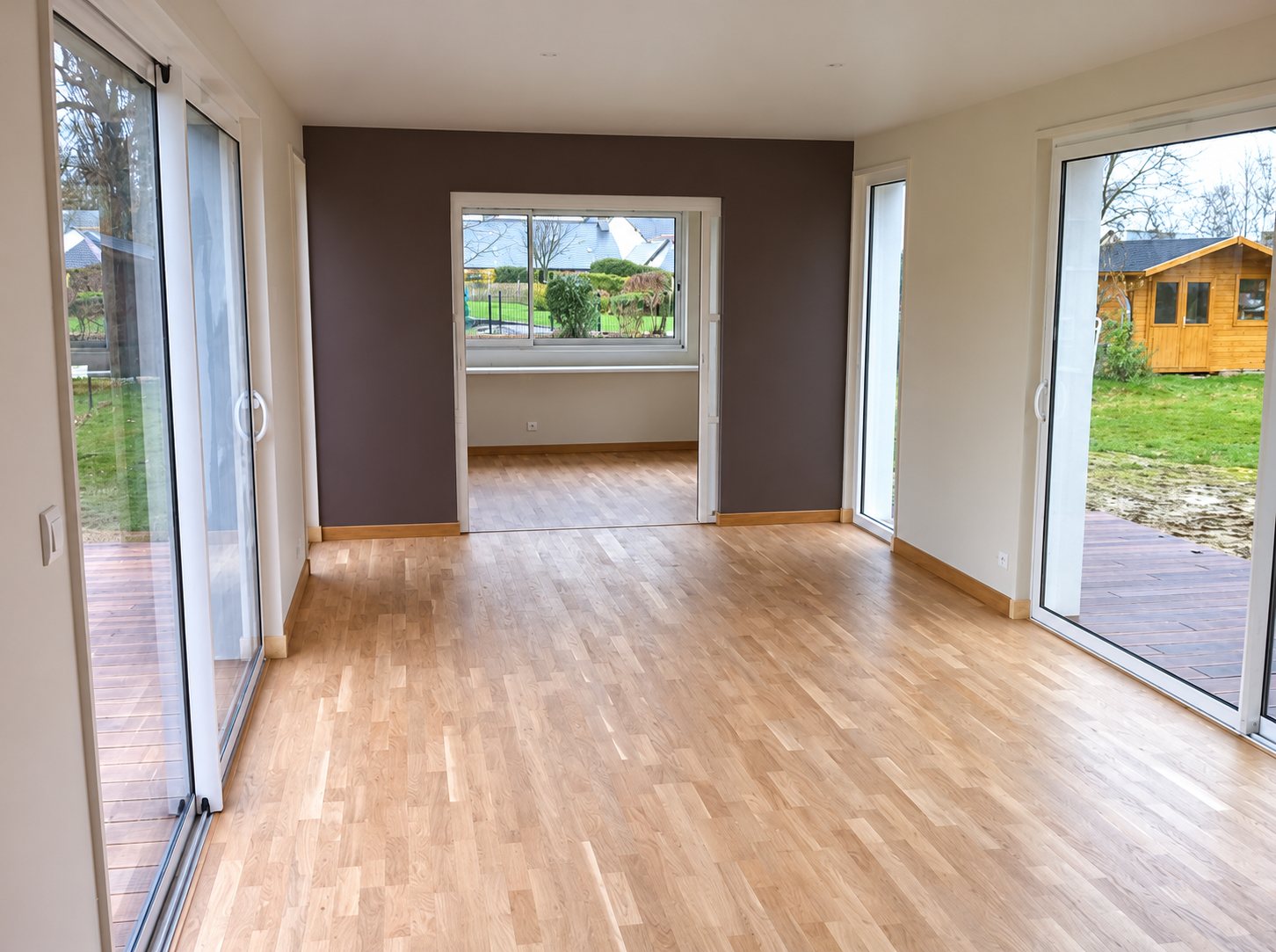 Pièce vide fraîchement rénovée avec un parquet en chêne clair, un mur prune et de grandes baies vitrées donnant sur un jardin et un chalet en bois.