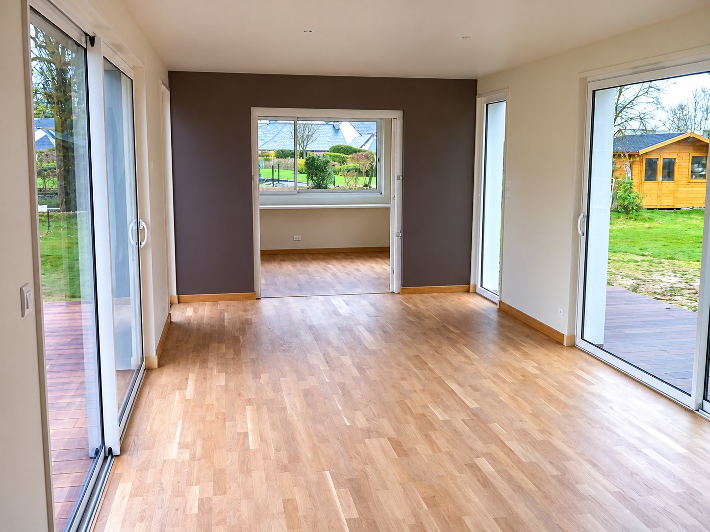 Pièce de vie vide avec parquet en chêne clair, mur prune et grandes baies vitrées coulissantes donnant sur un jardin.